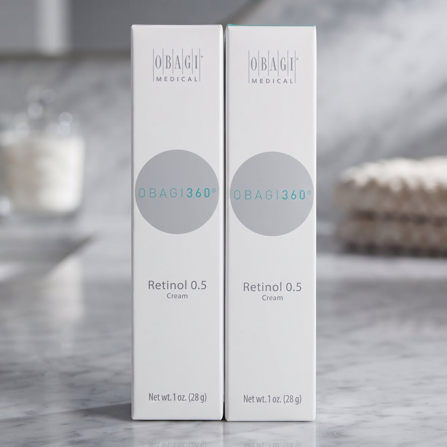 Two white OBAGI Medical OBAG1360 Retinol 0.5 Cream boxes standing upright on a light marble surface with a softly blurred bathroom background.