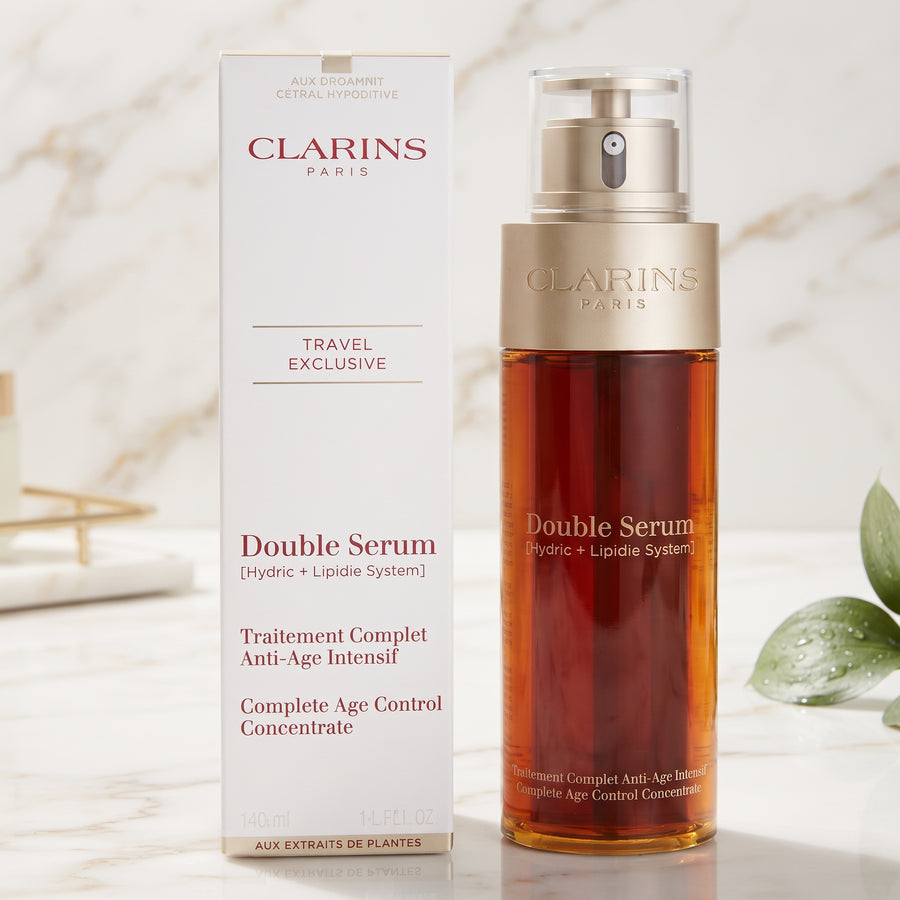 Clarins Paris Double Serum travel exclusive set, featuring a white box and a clear bottle filled with amber-colored serum, set on a light marble surface with soft green leaves in the background.