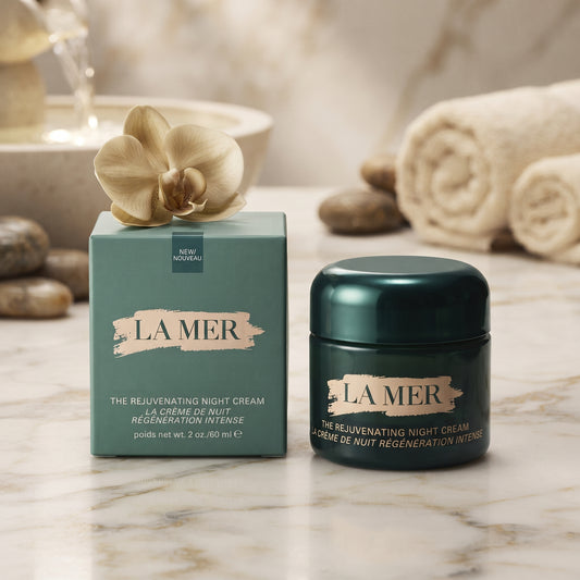 La Mer The Relivenating Night Cream shown with its teal outer box and dark teal jar on a marble surface, styled with rolled towels, stones, and a flower in a spa-like setting.