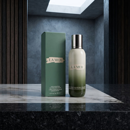 La Mer Hydrating Infused Treatment serum bottle with a silver cap standing next to its green box on a polished marble surface.