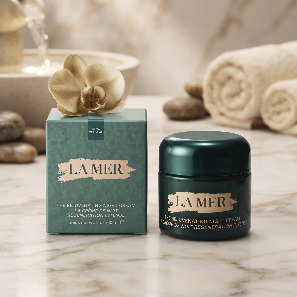 La Mer The Relivenating Night Cream shown with its teal outer box and dark teal jar on a marble surface, styled with rolled towels, stones, and a flower in a spa-like setting.